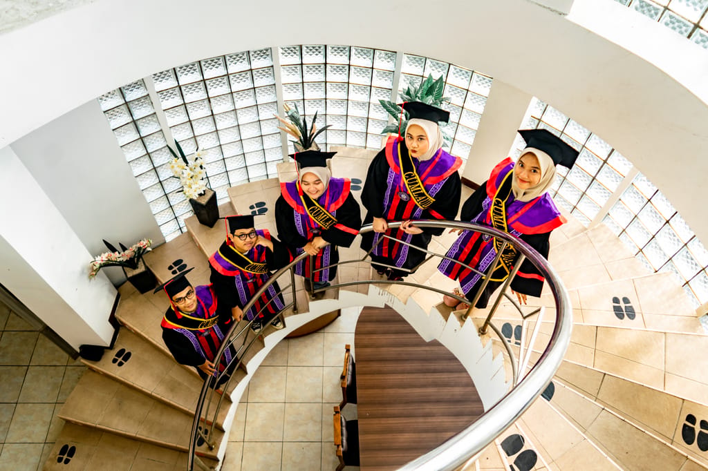 A group of graduates in colorful robes pose on a spiral staircase indoors.