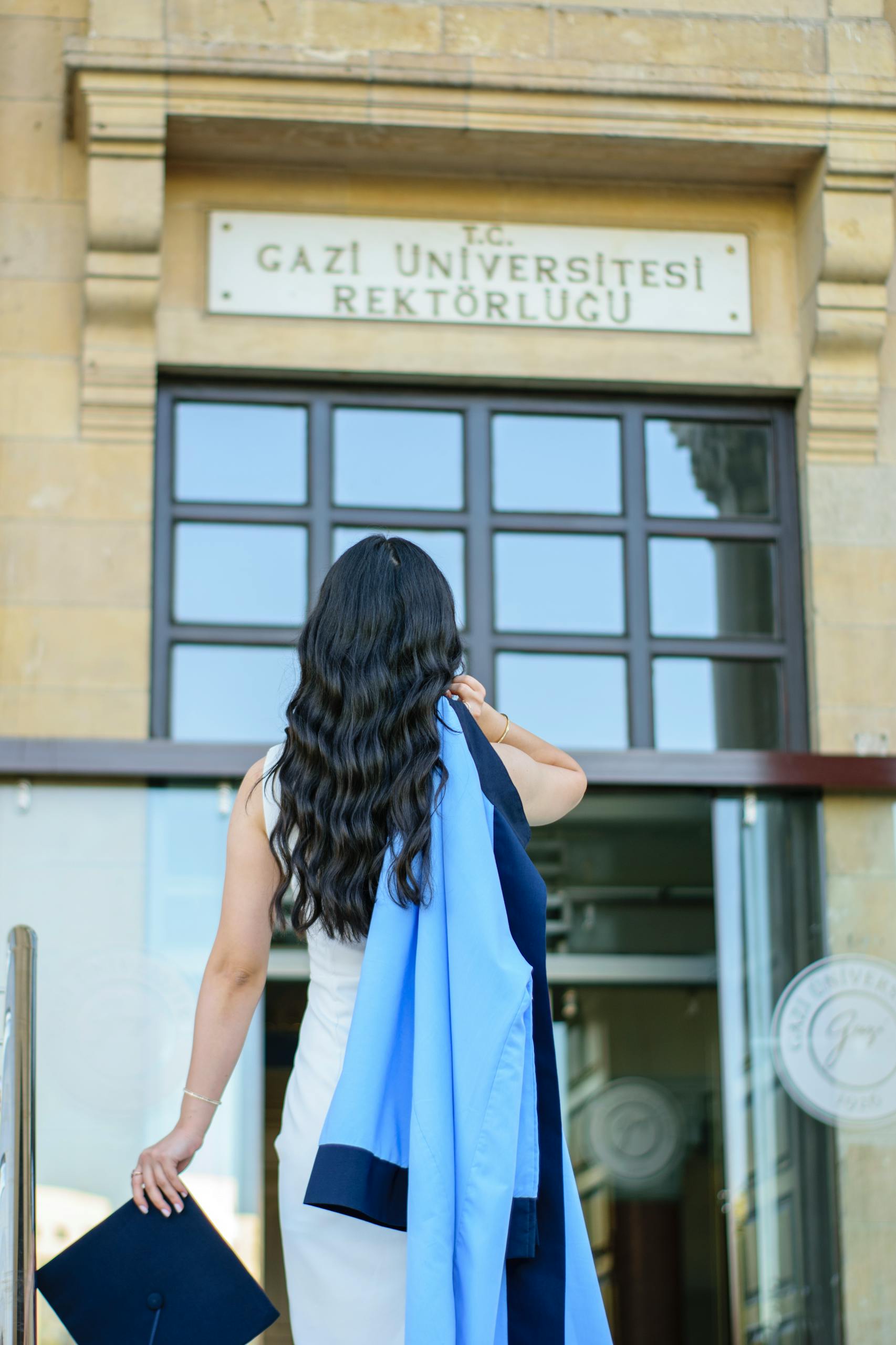 Brunette woman in graduation gown standing outside Gazi University entrance, holding cap.