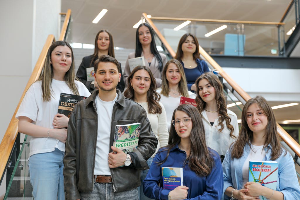 Diverse group of students smiling on stairs, holding educational books indoors.