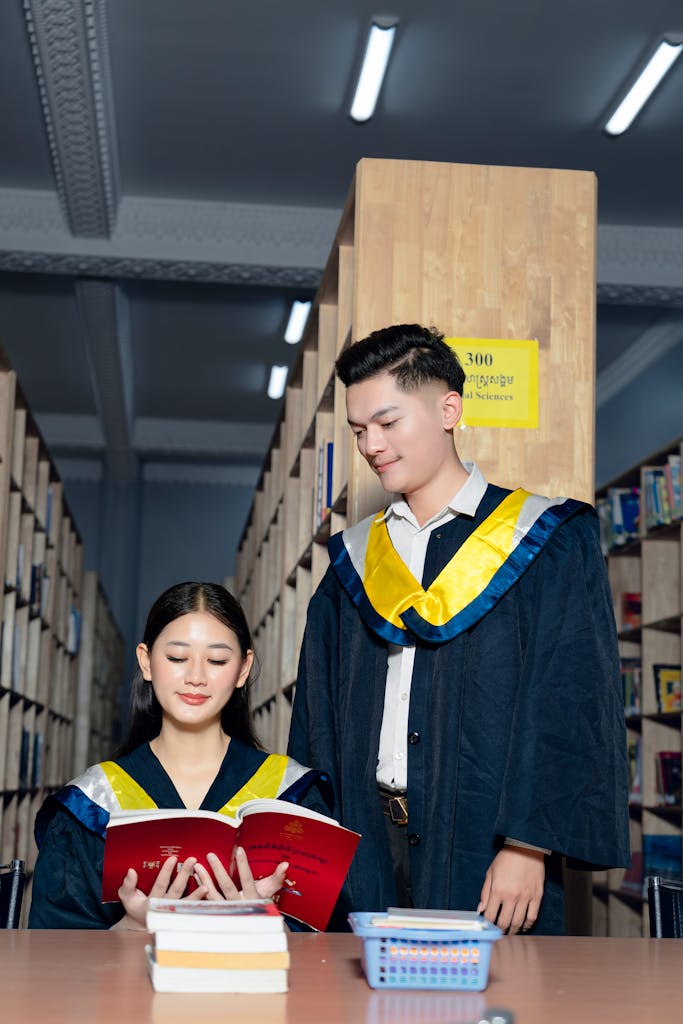 Two students in graduation gowns studying in a library setting.